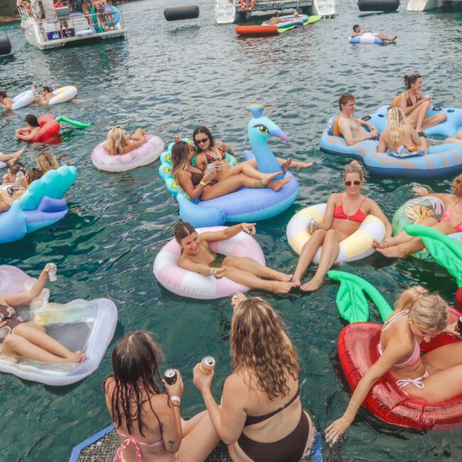 A group of people relax on colorful inflatable pool floats in the water, enjoying a sunny day. Many are wearing swimsuits, sunglasses, and holding drinks, with boats visible in the background.