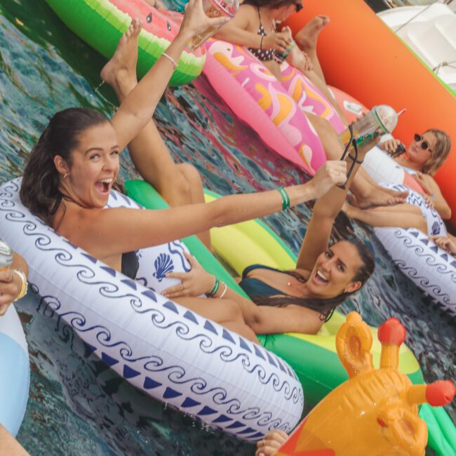 A group of people relax and celebrate on colorful pool floats in the water, raising drinks and smiling; the floats are shaped like animals, watermelon, and other playful designs.