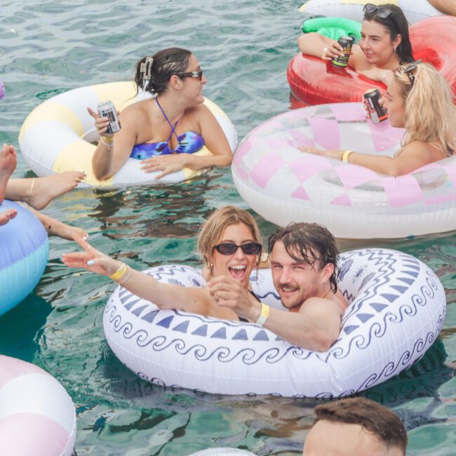 A group of people relax in colorful inflatable pool floats on water, laughing and holding drinks. The scene is lively and festive, with various float designs including a large blue swan and patterned rings.