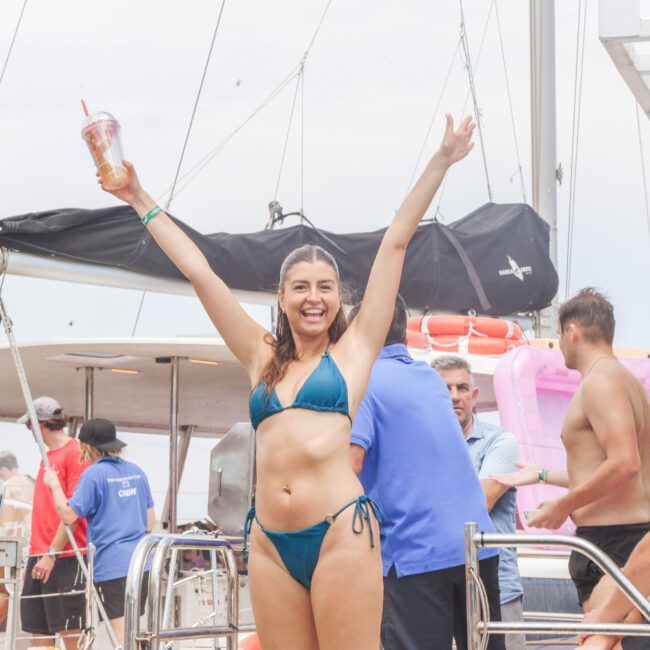 A smiling woman in a blue bikini raises her arms and holds a drink on a boat, surrounded by other people enjoying a sunny day.