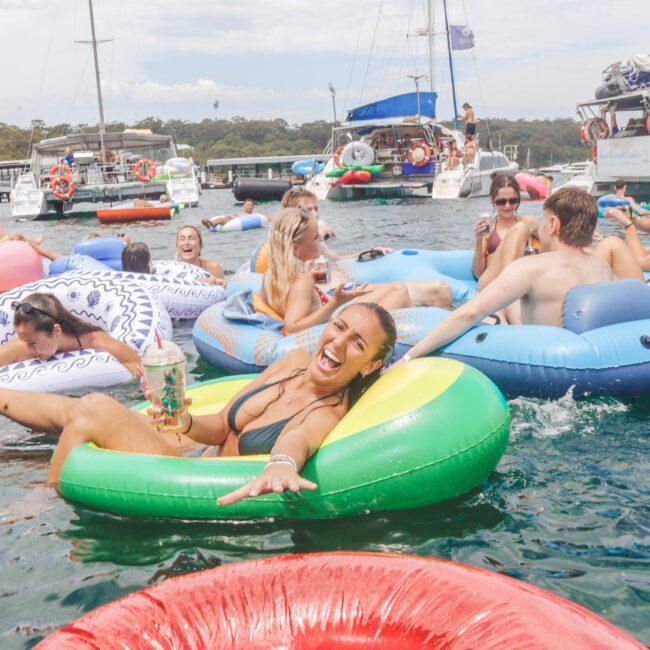 A group of people are relaxing on colorful inflatable floats in the water near boats. A smiling woman in a green float holds a drink and poses for the camera, surrounded by others enjoying the sunny day.