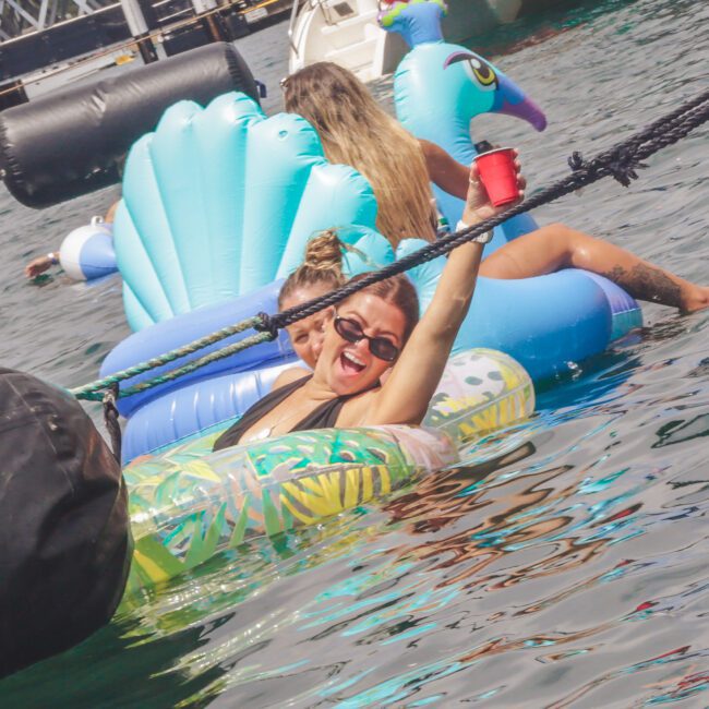 Two women relax on colorful inflatable pool floats in a body of water. The woman in front, smiling and wearing sunglasses, holds up a red cup. In the background is a dock and more inflatables.