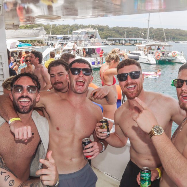 A group of young men in swimwear and sunglasses pose and smile together on a boat during a lively party, holding drinks, with other people and boats visible in the background on the water.