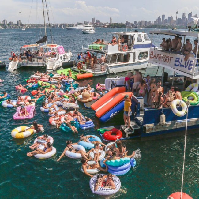 A lively boat party with many people on inflatable floats in the water, gathered around docked boats on a sunny day with city buildings and more boats visible in the background.