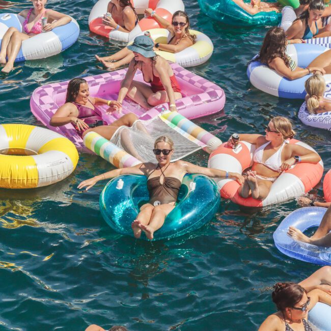 A group of people in swimsuits relax on colorful inflatable pool floats in clear blue water, enjoying drinks and sunshine. The scene is lively and summery, with everyone smiling and having fun.