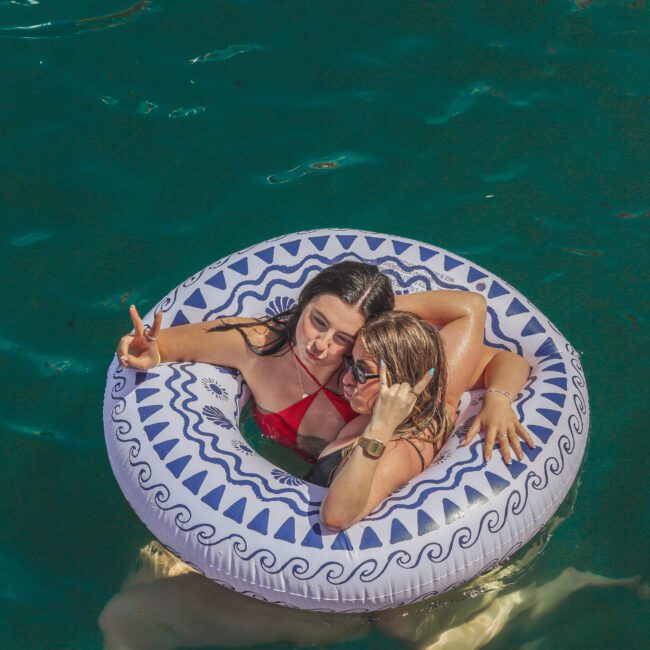 Two women relax on a patterned inflatable pool float in clear turquoise water. One woman flashes a peace sign while they both smile and pose for a selfie. Sunlight reflects on the water’s surface.