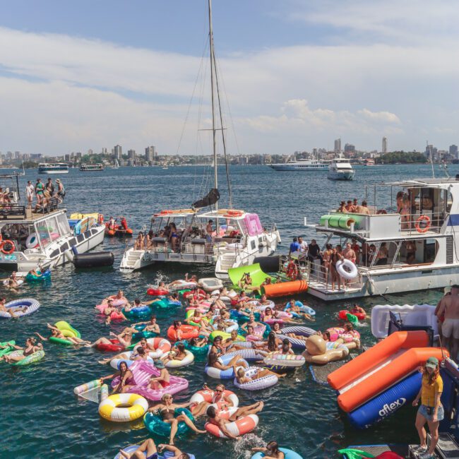 Dozens of people on colorful inflatables relax and socialize in the water, surrounded by anchored boats on a sunny day with a city skyline visible in the background.