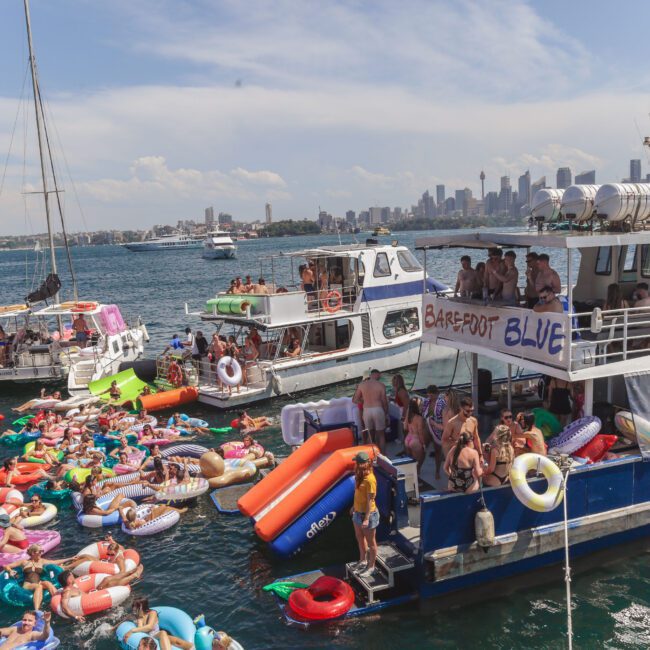 A group of people party on boats and colorful pool floats in the water near a city skyline under a partly cloudy sky. The boat “Barefoot Blue” is docked beside others with people dancing and sunbathing.