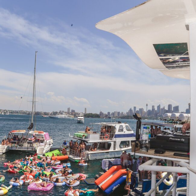 A DJ plays music on a boat overlooking a crowded party on the water, where people on inflatable floats and boats enjoy the sunny day with a city skyline in the background.