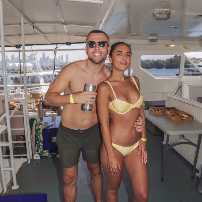 A smiling man in sunglasses and swim shorts stands next to a woman in a yellow bikini on a boat. The man holds a tumbler, and the woman has her hand on his waist. Food and drinks are visible on tables in the background.