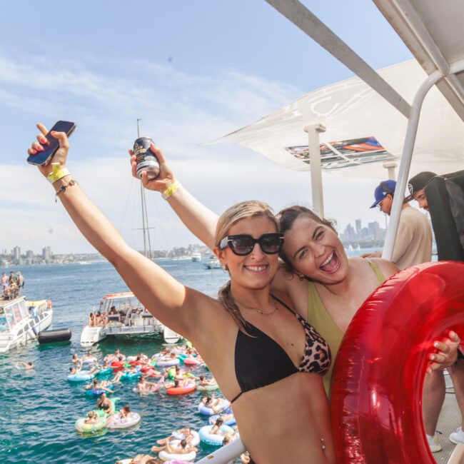 Two young women in swimsuits smile and pose on a boat, one holding a drink and the other a red pool float. Behind them, people on boats and inflatables enjoy a sunny day on the water, with a city skyline in the distance.