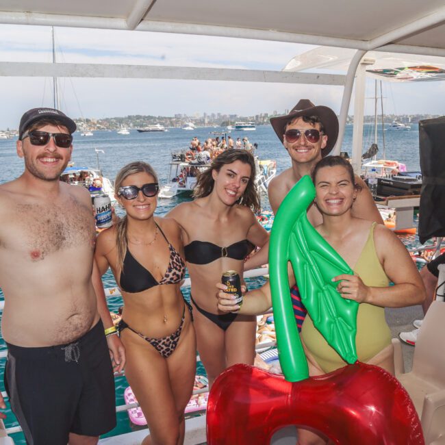 Five people in swimsuits smile for a photo on a boat. The group includes three men and two women; one person holds an inflatable cherry. The background shows water, boats, and people enjoying a sunny day.