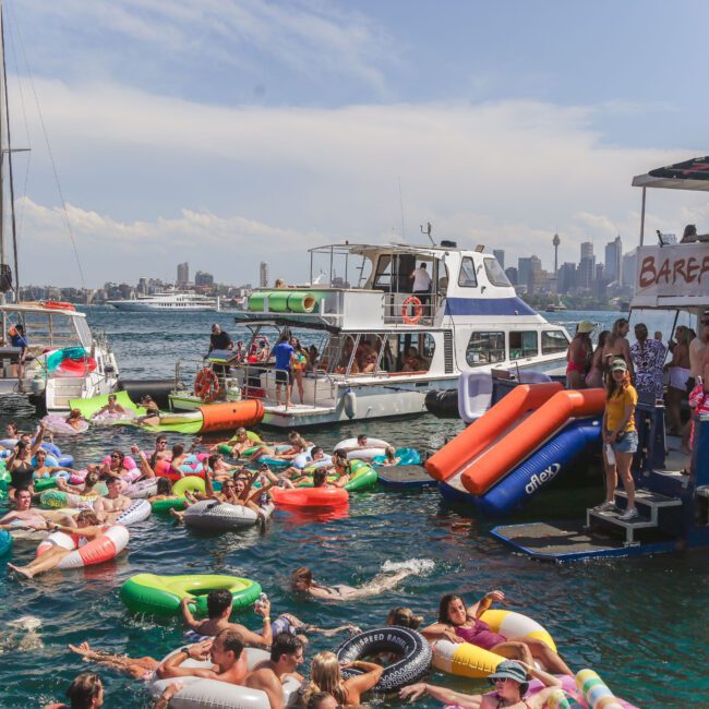 A large group of people on colorful inflatables enjoy a lively boat party on the water near several anchored boats, with a city skyline visible in the background under a partly cloudy sky.