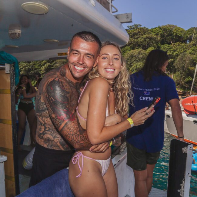 A smiling man and woman in swimwear stand hugging on a boat, with water, trees, and other people visible in the background. The man has tattoos on his arms, and the woman is holding a phone.