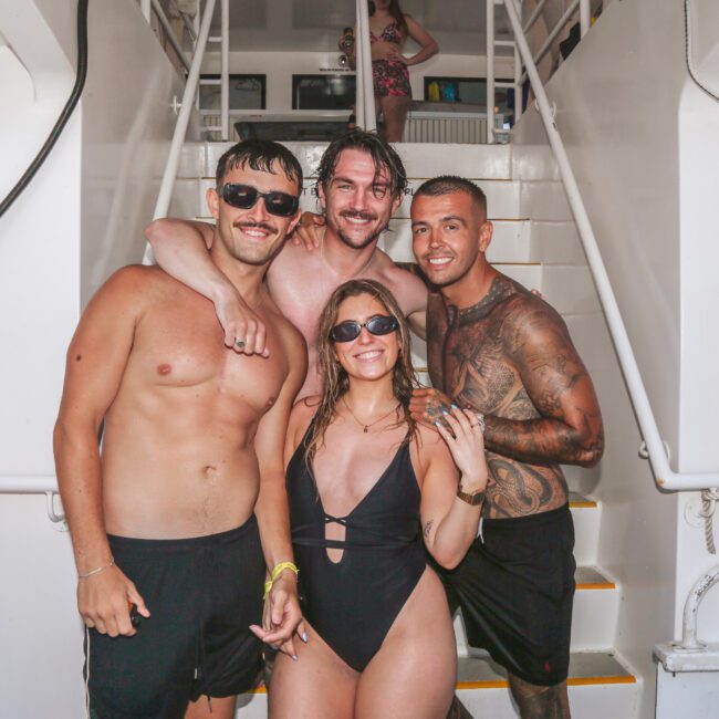 Four young adults in swimwear, smiling and posing together on the deck of a boat. Three men stand behind a woman in a black swimsuit. The setting is bright and casual, with white staircases in the background.