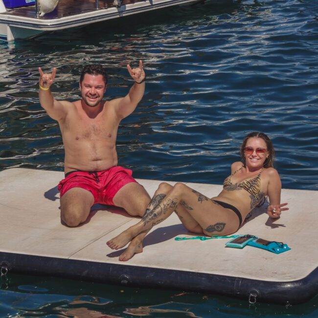 A man and a woman relax on a floating mat in the water. The man, wearing red shorts, sits with arms raised, smiling. The woman, in a patterned bikini, lies on her side, also smiling, with a boat visible in the background.
