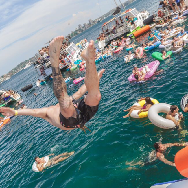 A person mid-air after jumping into the sea, surrounded by people floating on colorful inflatables at a lively waterfront event with boats and a crowded pier in the background.