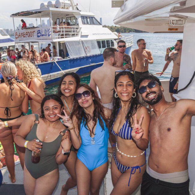 A group of smiling friends in swimsuits pose on a boat deck, flashing peace signs. Other people socialize and swim nearby, with boats and clear blue water in the background on a sunny day.