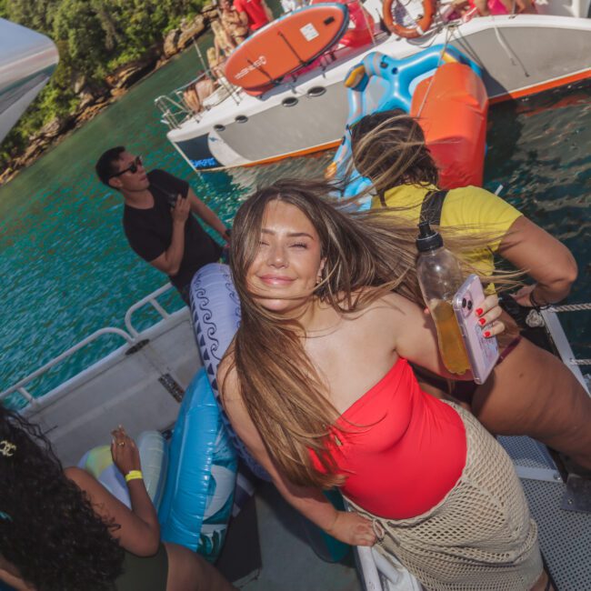 A woman in a red swimsuit and beige cover-up smiles at the camera on a boat, holding a drink. Other people are gathered nearby, with water and another boat in the background on a sunny day.