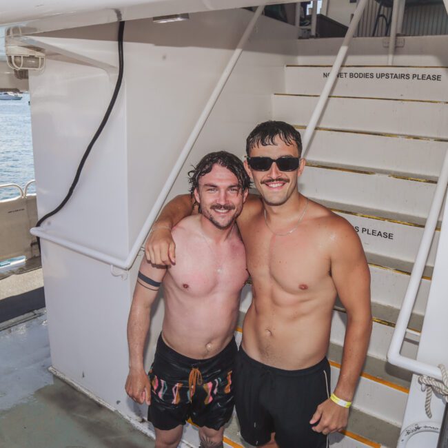 Two smiling men in swim trunks stand arm-in-arm on a boat deck, with other people in swimwear in the background. Stairs with "GET BODIES UPSTAIRS PLEASE" written on them are visible behind them.