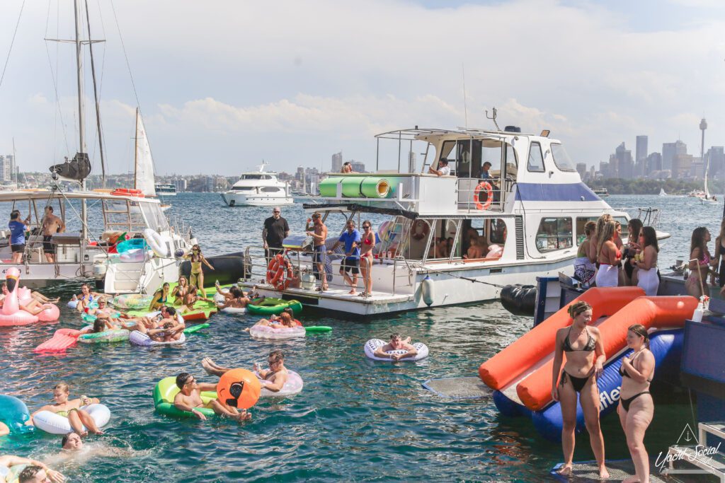 A lively boat party with people swimming and lounging on colorful inflatables in the water, others relaxing on boats, and a city skyline visible in the background under a partly cloudy sky.