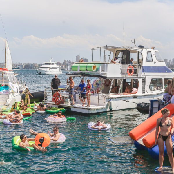 A lively boat party with people swimming and lounging on colorful inflatables in the water, others relaxing on boats, and a city skyline visible in the background under a partly cloudy sky.