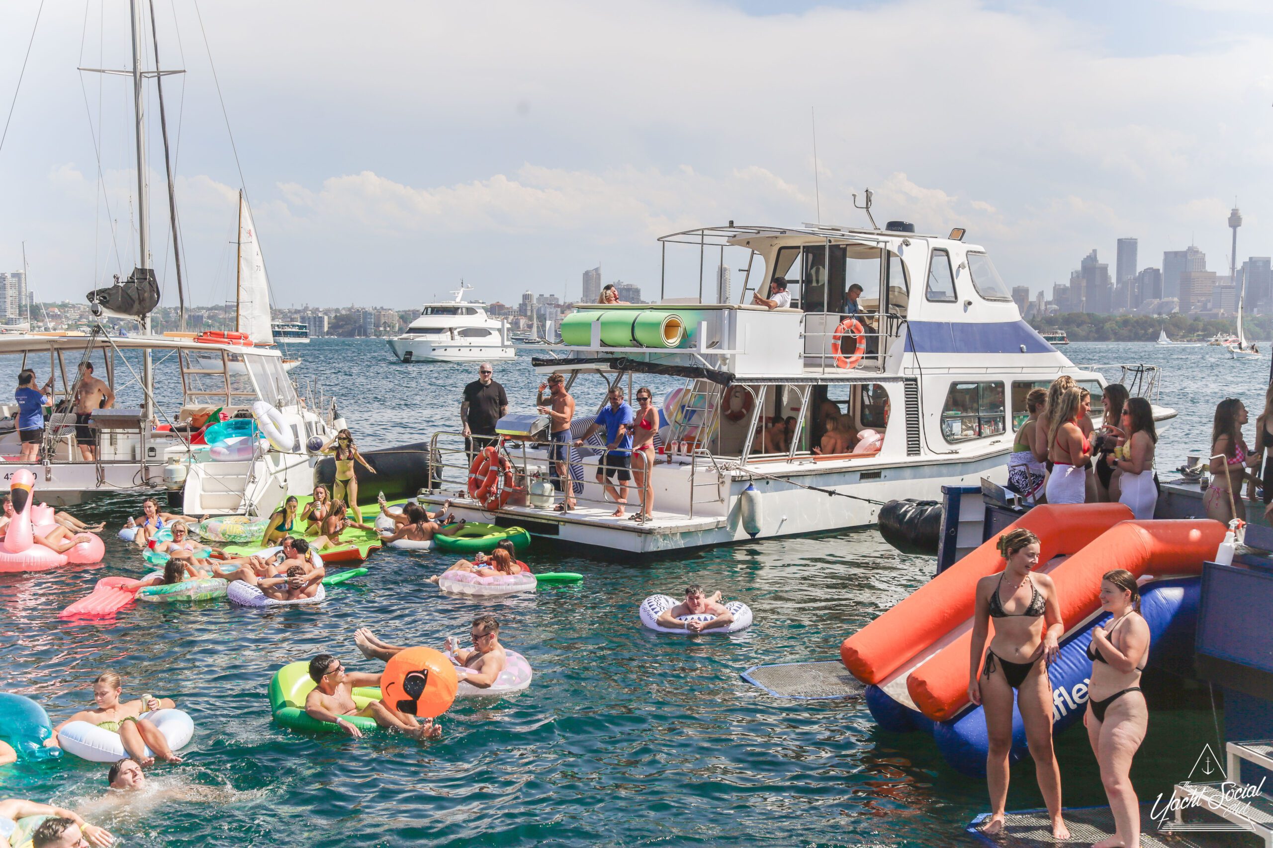 A lively boat party with people swimming and lounging on colorful inflatables in the water, others relaxing on boats, and a city skyline visible in the background under a partly cloudy sky.