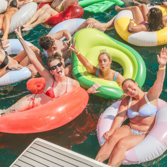 A group of young adults relax in colorful pool floats on a sunny day, smiling, waving, and making playful faces in the water near a dock.