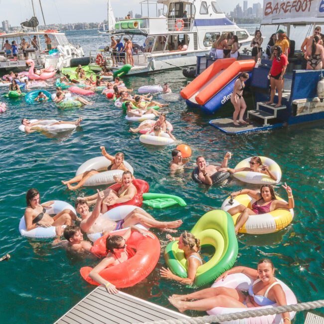 A lively group of people relax on colorful pool floats in the water near docked boats during a sunny party; others stand and socialize on the boats in the background.