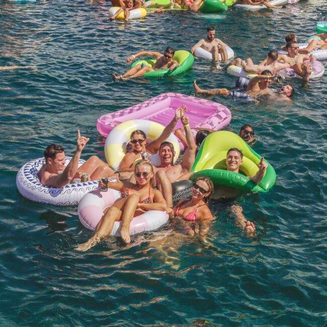 A group of people on colorful pool floats smile and pose for a photo in a crowded, sunny outdoor pool, surrounded by others relaxing and enjoying the water on inflatables.