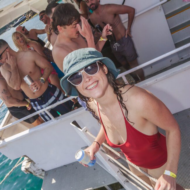 A woman in a red swimsuit, sunglasses, and a blue bucket hat smiles at the camera while holding a drink on a boat. Several people in swimwear are socializing in the background. The boat is on the water.