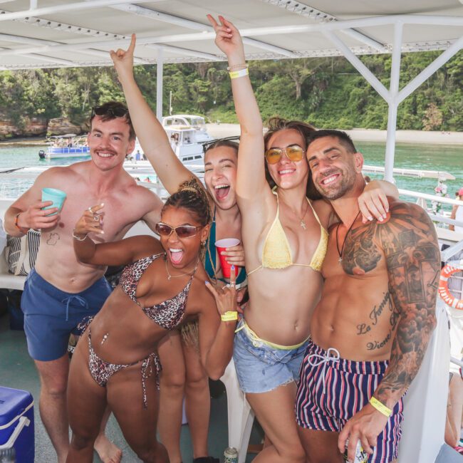 Five friends in swimsuits smile and pose energetically on a boat, holding drinks. Green water and trees are visible in the background, with other boats nearby. It appears to be a lively summer outing or party.