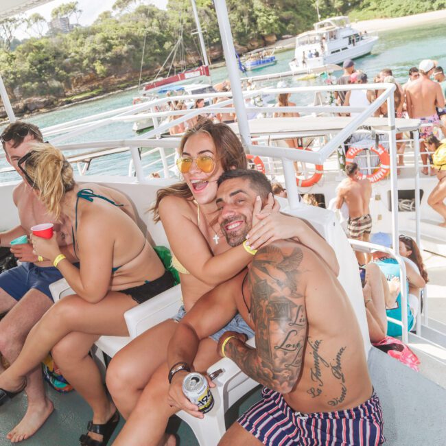 A smiling man and woman in swimwear sit closely and hug on a boat, surrounded by other people enjoying a sunny day. The background shows calm water, boats, and lush green trees.