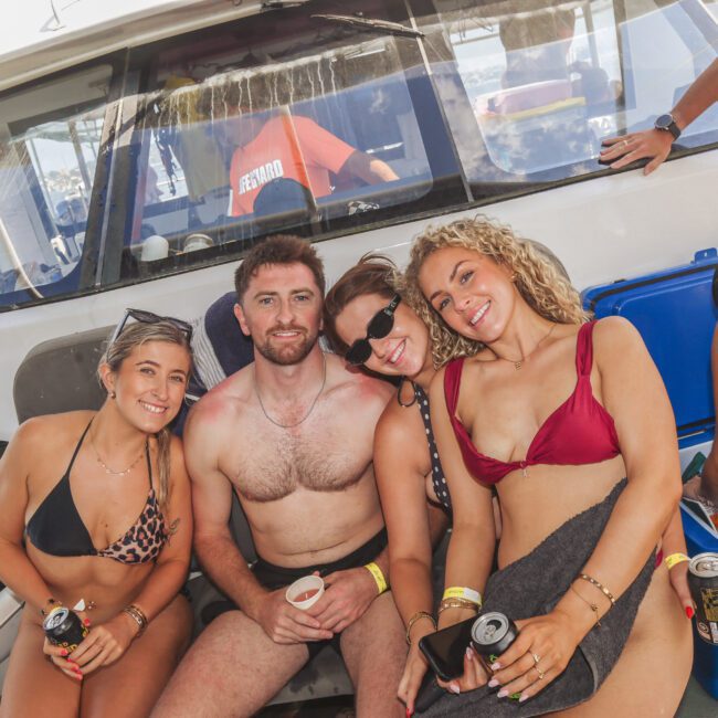 Four people in swimsuits sit close together on a boat, smiling and holding drinks. There are coolers nearby and another person in the background. The atmosphere appears relaxed and sunny.
