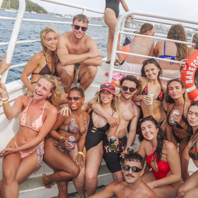 A group of people in swimsuits smile and pose together on a boat deck, holding drinks. The sea and coastline are visible in the background, and a lifebuoy labeled "Barefoot Spirit" hangs on the railing.