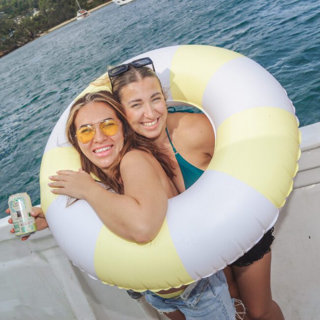 Two smiling women in swimsuits hug and pose with a large yellow and white inflatable ring on a boat. One holds a can, and blue water with trees is visible in the background. Part of another person is seen on the right.