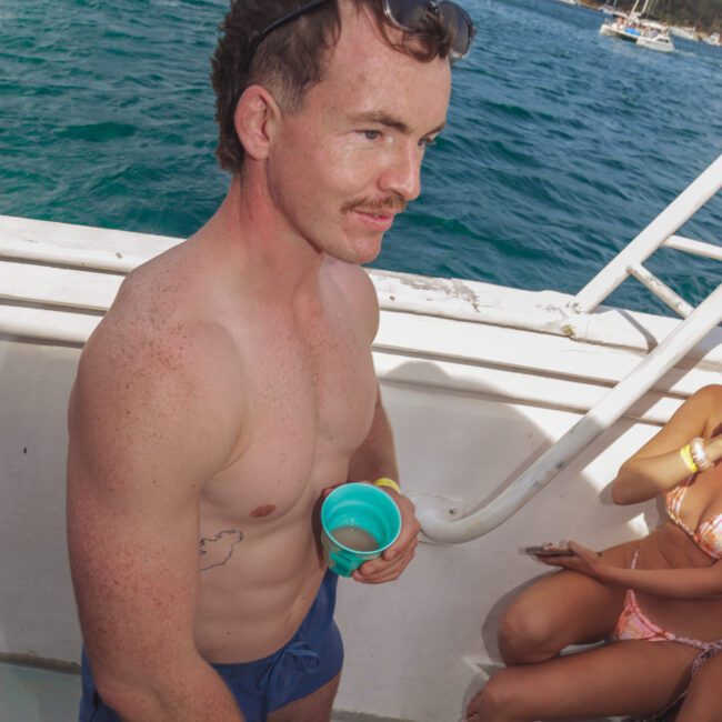 A shirtless man with a mustache, sunglasses on his head, and blue swim trunks holds a green cup on a boat. The ocean and anchored boats are visible in the background. Other people are partially visible nearby.