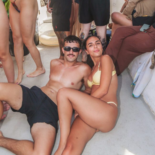 A man and woman in swimsuits relax on the floor of a crowded boat, with others standing and socializing around them. The man wears sunglasses and black shorts; the woman wears a yellow bikini.