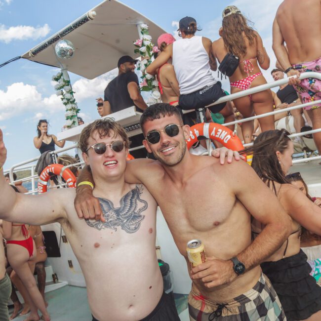 Two young men in swimwear and sunglasses pose, smiling with arms around each other, at a lively boat party full of people dancing and socializing in the sun.