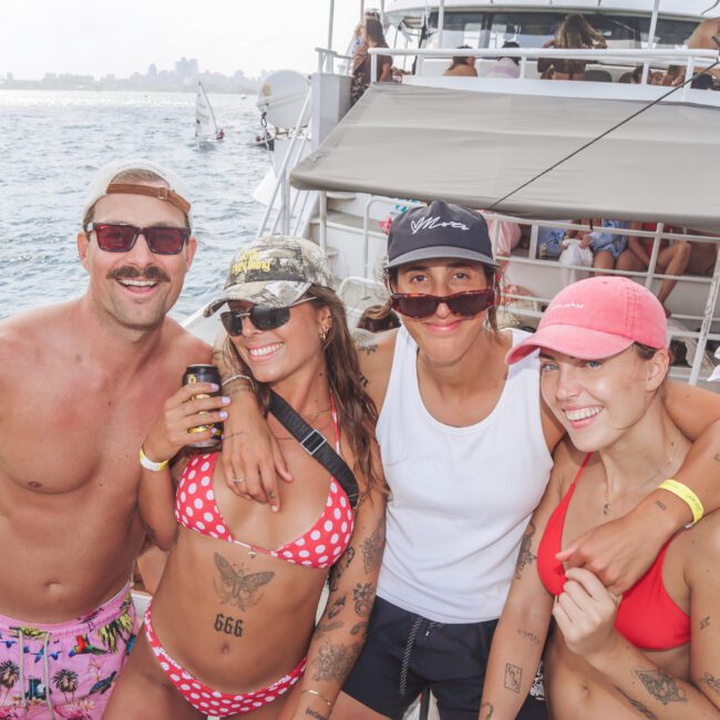 Four smiling people in swimwear pose together on a boat. The group is enjoying a sunny day, with water and other boats visible in the background. The atmosphere is lively and festive.