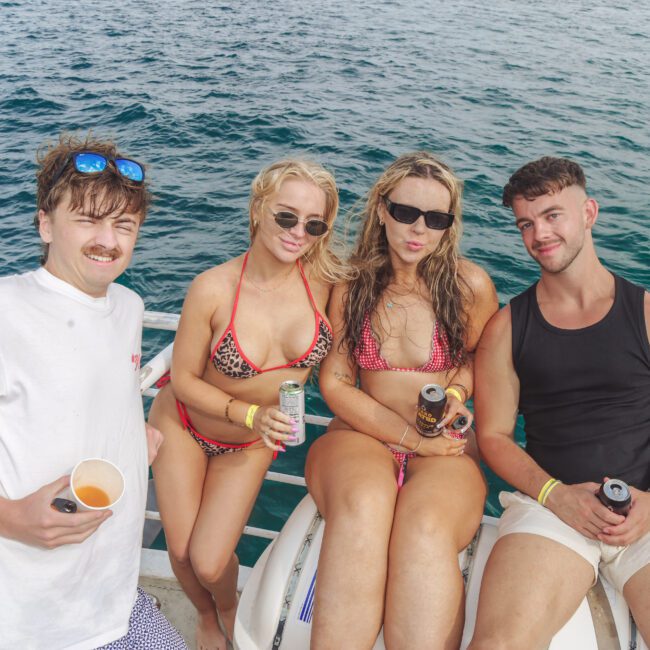 Four young adults, two men and two women, sit together on the railing of a boat, smiling and holding drinks. The background shows clear blue ocean water.