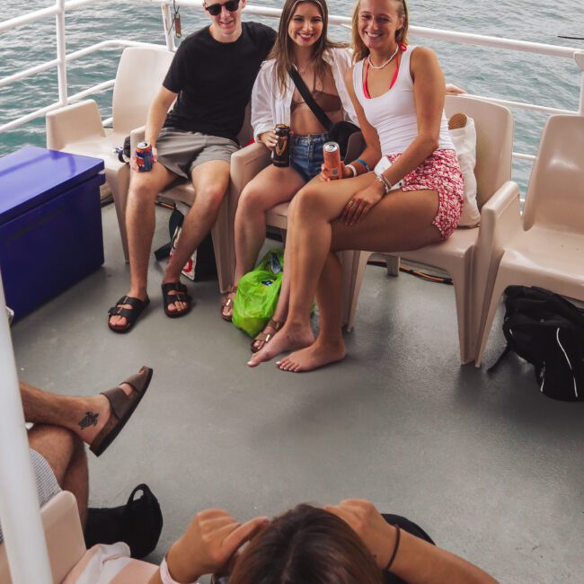 Three people sit smiling on a boat with drinks in hand, while another person sits in the foreground facing them. The boat is on the water and the group appears relaxed and happy.