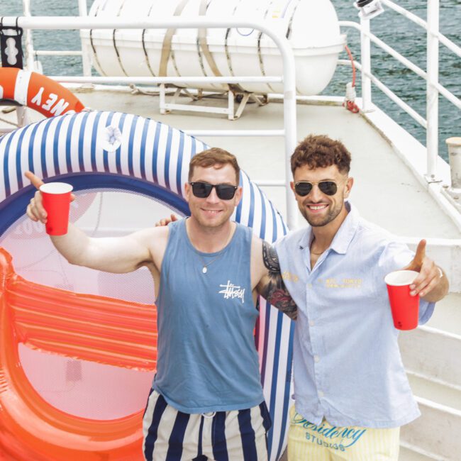 Two men smiling and holding red cups pose on a boat next to a striped inflatable pool float, with life preservers and water in the background. Both wear sunglasses and summer clothes.