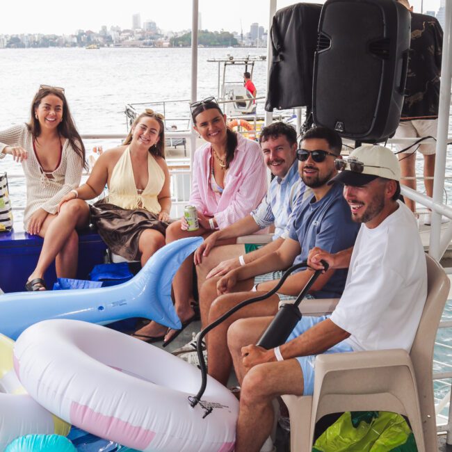 Six people sit and smile together on a boat, surrounded by inflatable pool toys and a speaker. The city skyline and water are visible in the background, creating a relaxed, cheerful atmosphere.