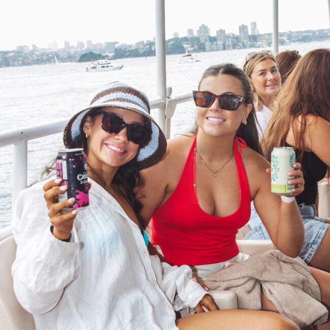 Two women sitting on a boat, smiling at the camera while holding canned drinks. One wears a white shirt and sunhat, the other a red swimsuit and sunglasses. Other people are in the background, with water and cityscape behind them.