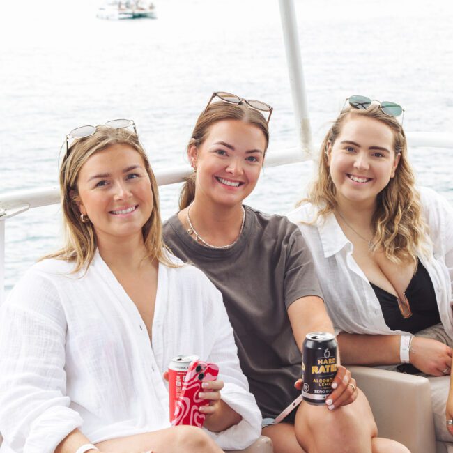 Three women sitting on a boat, smiling at the camera. They are wearing casual summer clothes and sunglasses, holding canned drinks. The water is visible in the background.