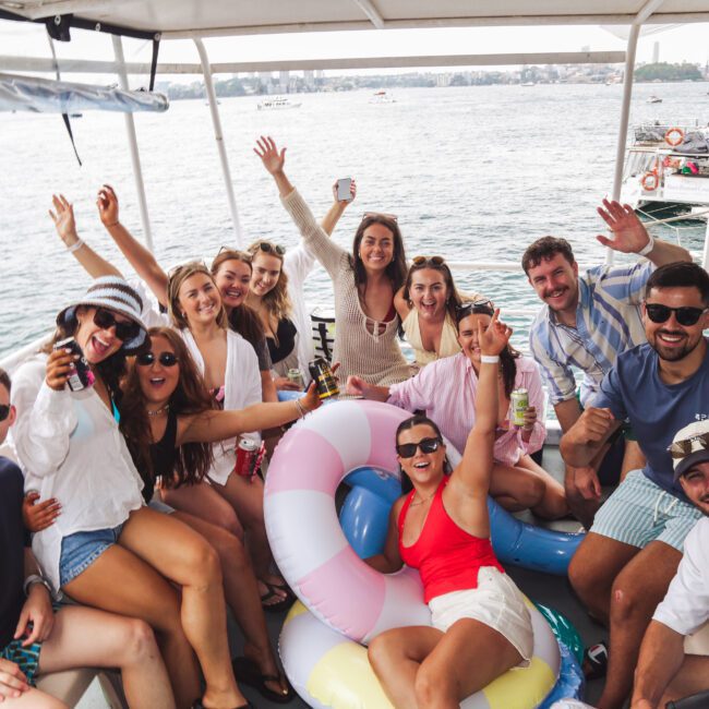 A group of friends on a boat smile and wave at the camera, holding drinks and wearing casual summer clothes. They are surrounded by inflatables and water, enjoying a lively day on the lake.