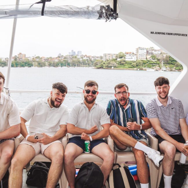 Five young men sit on a boat bench, smiling and laughing together. They are casually dressed in shorts and shirts, holding drinks, with water and city buildings visible in the background.