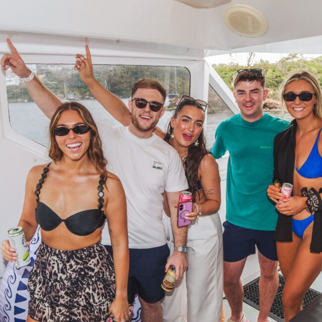 Five smiling young adults, wearing summer outfits and sunglasses, pose together on a boat holding drinks. Lush greenery and water are visible through the boat's window in the background.