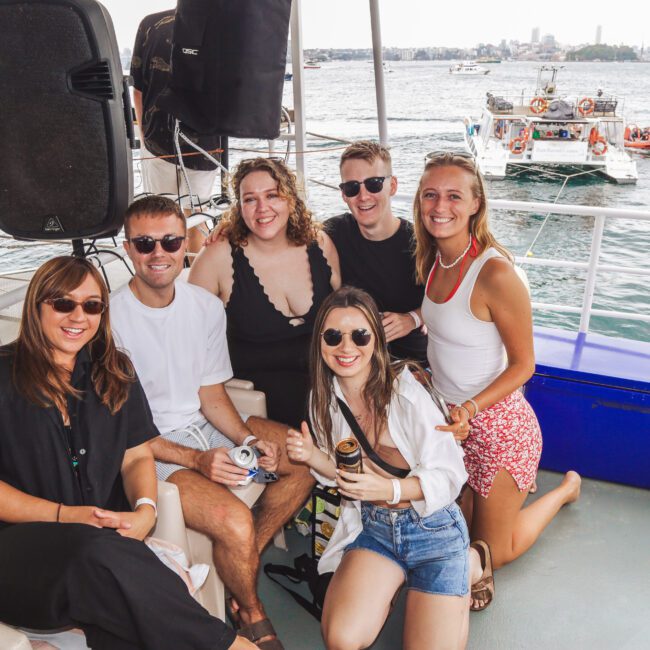 A group of six smiling people pose together on a boat, with water, boats, and a city skyline visible in the background. It’s a sunny day and everyone is dressed in casual summer clothing.